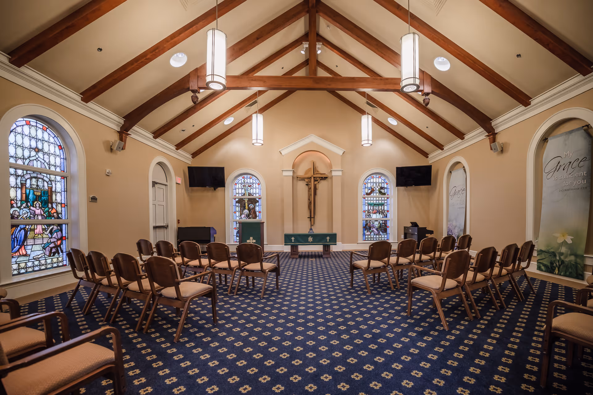 Interior view of a chapel with wooden beams on the ceiling, stained glass windows, a cross mounted on the wall behind an altar, and rows of chairs arranged facing the altar. The room has patterned carpet and hanging pendant lights.
