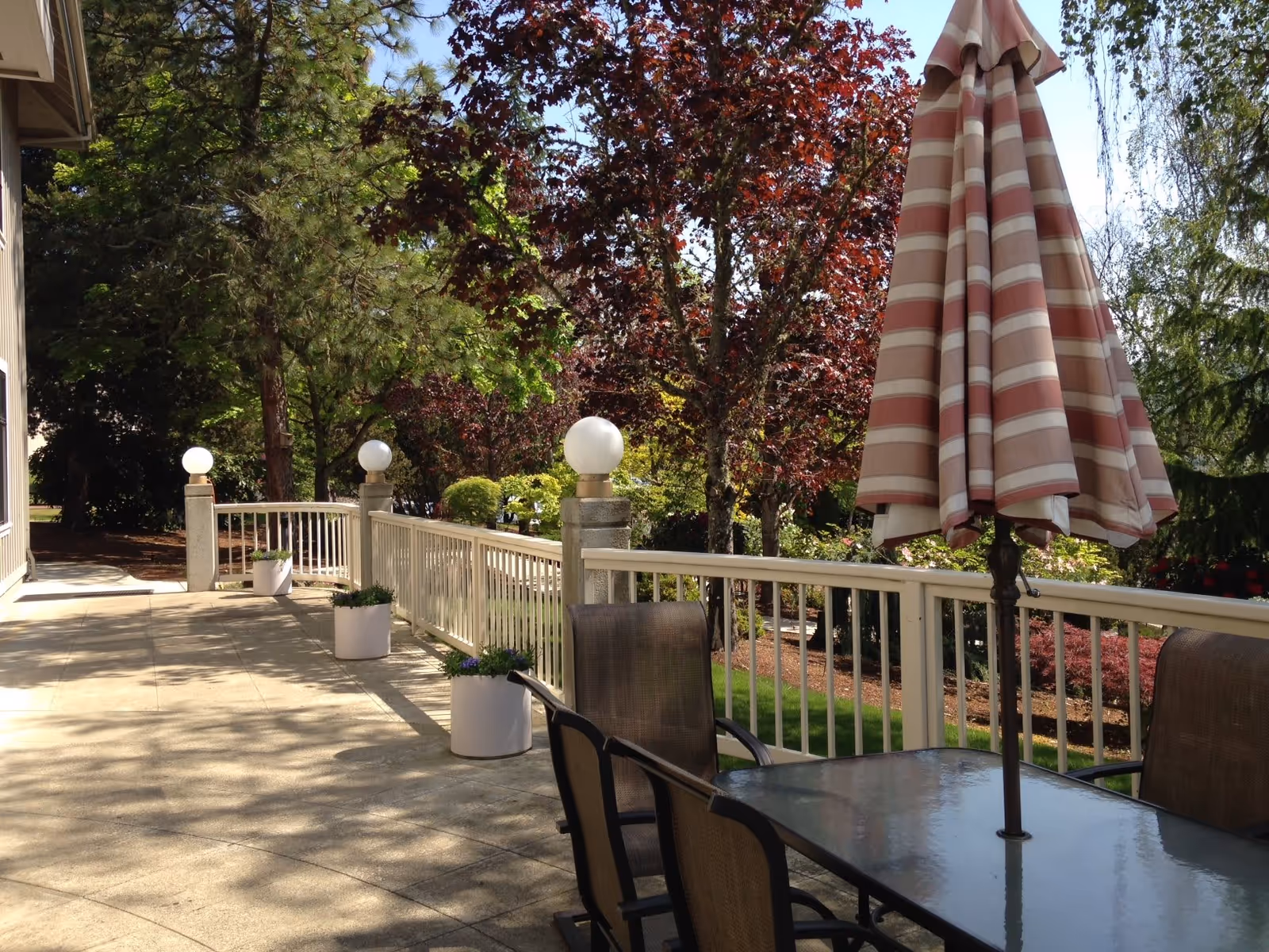 Outdoor patio area with a glass table and four chairs, a closed striped umbrella, white railing, potted plants, and trees with green and red leaves in the background.