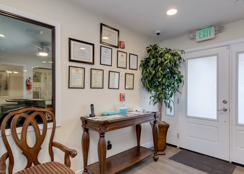 Interior view of a reception or waiting area in West Drive Assisted Living and Memory Care featuring a wooden chair with a striped cushion, a wooden console table with documents, a phone, and medical supplies on top, a large potted plant, framed certificates on the wall, and a white door with frosted glass panels and an exit sign above.