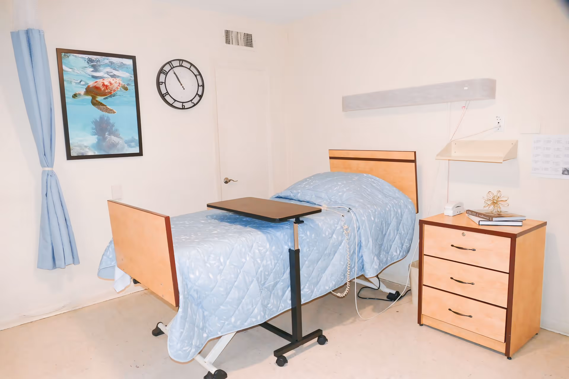 A simple bedroom in a care facility with a single bed covered in a light blue quilt, a small overbed table, a wooden nightstand with three drawers, a clock on the wall, a framed picture of a sea turtle underwater, and light blue curtains on the window.