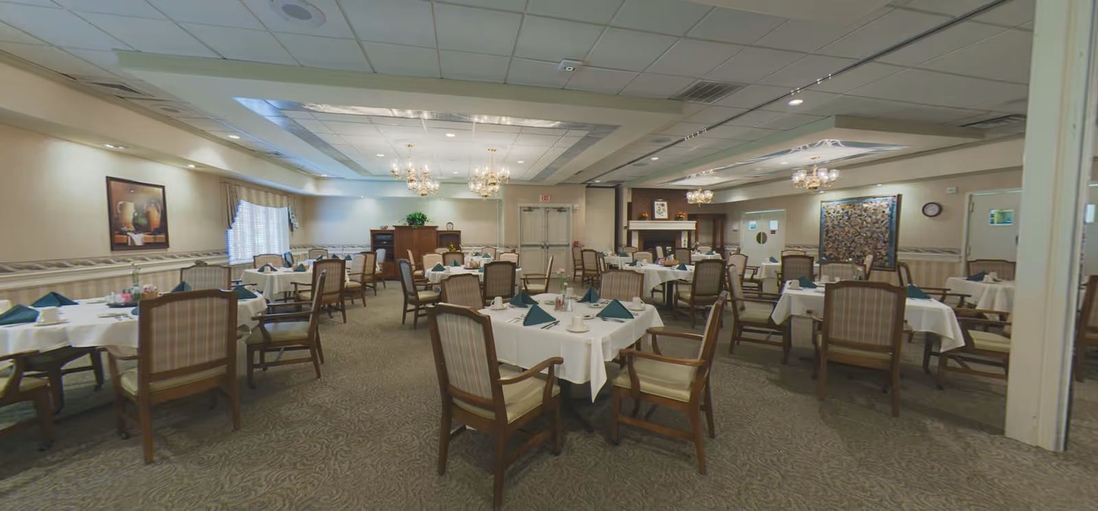 A spacious dining room with multiple round tables covered with white tablecloths, each set with green folded napkins, cups, and utensils. The room features wooden chairs with cushioned seats and backs, chandeliers hanging from the ceiling, a carpeted floor, and decorative wall art. There is a fireplace in the background and a cabinet with plants and decorative items.