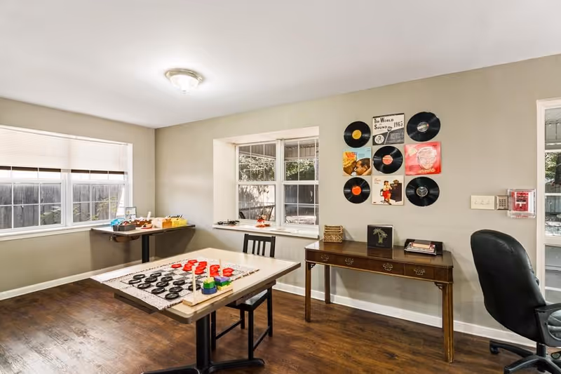 A bright room with large windows letting in natural light. There is a table in the center with a checkers game set up, a chair, and a wooden desk against the wall with books and decorative items. The wall above the desk is decorated with vinyl records and album covers. The floor is wooden, and there is an office chair near the desk.
