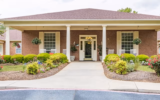 Front exterior view of a single-story brick building with a covered entrance supported by two white columns. There are white rocking chairs on the porch, hanging flower baskets, and well-maintained landscaping with bushes and flowers along the walkway leading to the entrance.