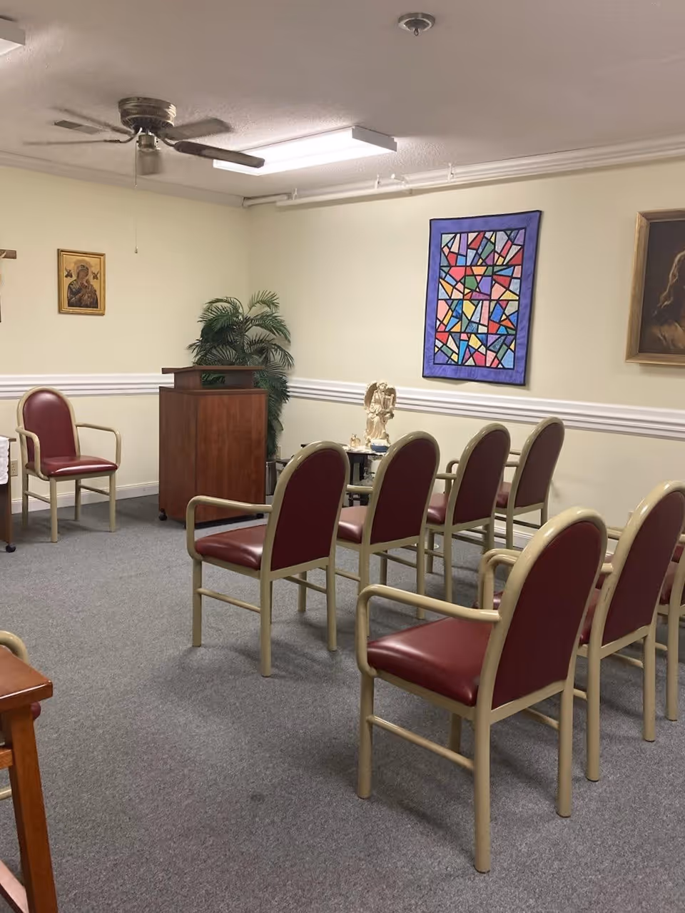 A small room set up with several chairs arranged in rows facing a wooden podium. The chairs have beige frames and red cushions. The walls are decorated with religious artwork, including a colorful stained glass style piece and framed portraits. A potted plant is placed near the podium, and the room has a ceiling fan and fluorescent lighting.