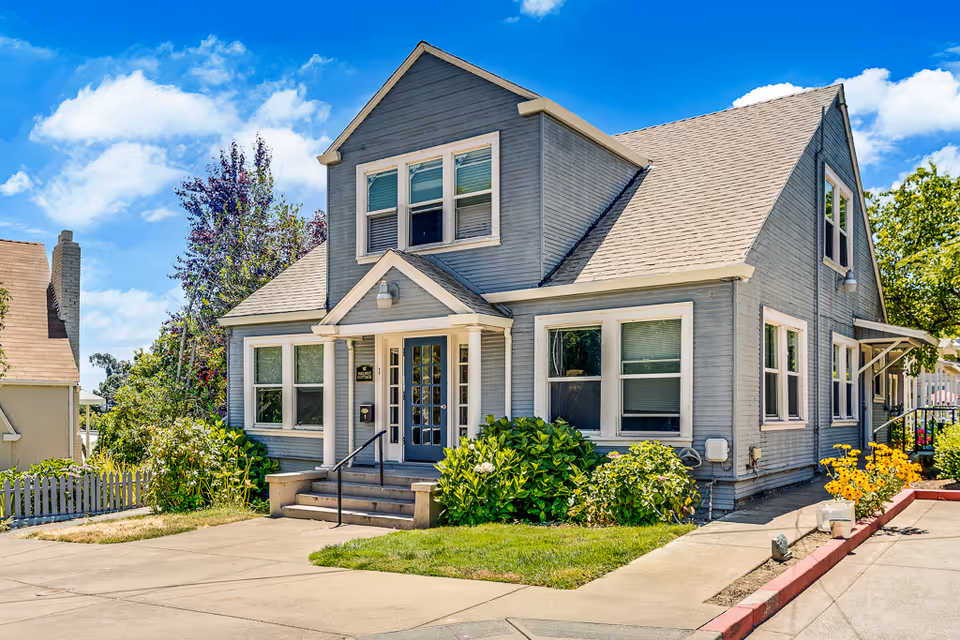 Front exterior of a gray two-story house with a small porch, surrounding shrubs, and a paved driveway under a blue sky.