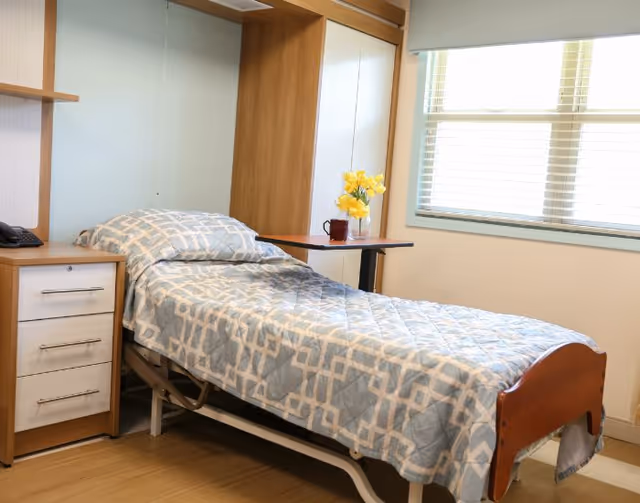 A bright senior-care bedroom with a single adjustable bed, bedside drawers, an overbed table with flowers, and a window with blinds.
