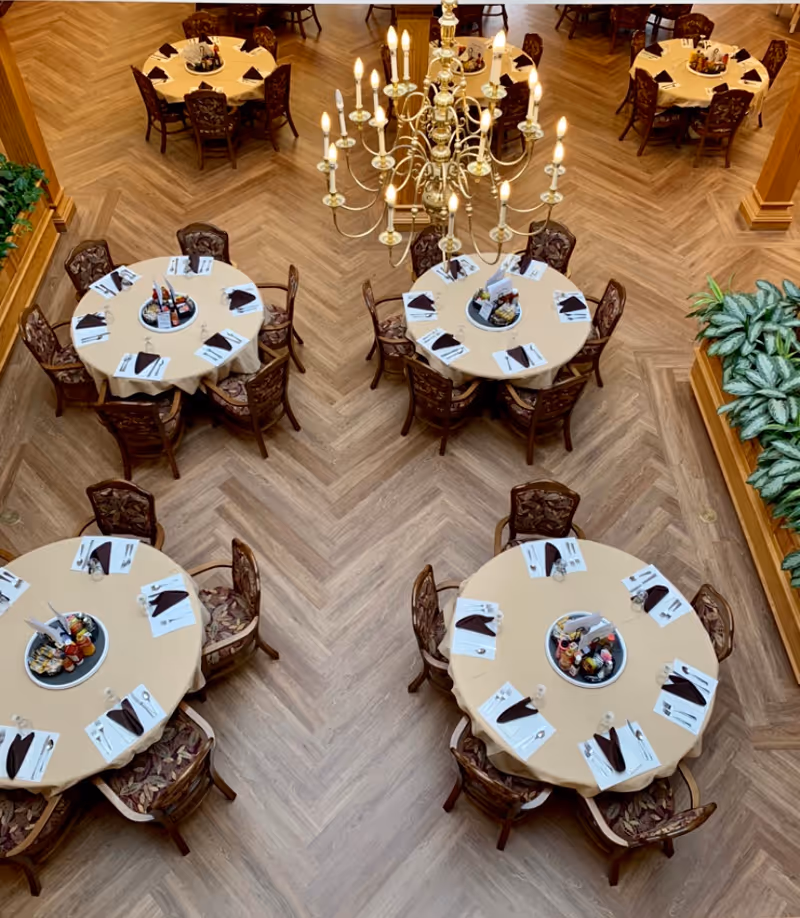 Overhead view of a dining room with round tables covered in beige tablecloths, each set with napkins, utensils, and condiment trays. Ornate wooden chairs with patterned cushions surround the tables. A large, elegant chandelier with multiple lit candles hangs above the center of the room. The floor has a herringbone wood pattern and there are green plants along the edges.