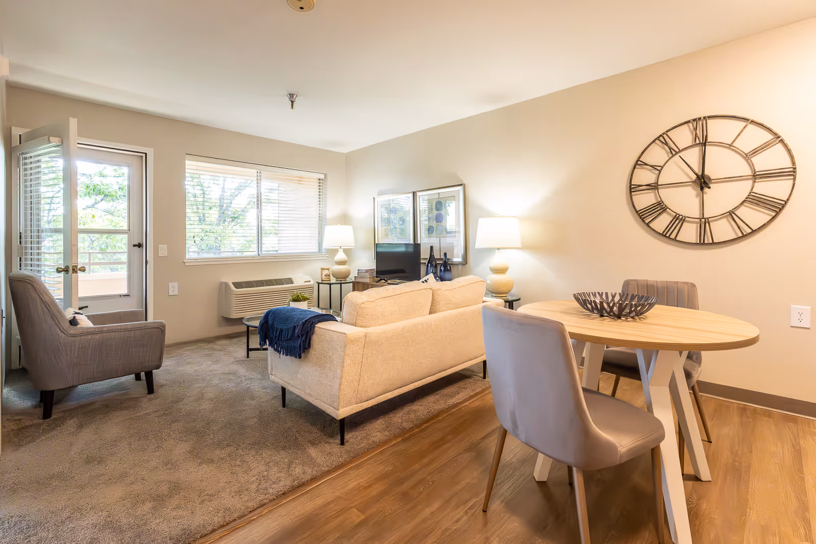 Bright furnished living and dining area with a sofa, armchairs, round dining table, large wall clock, window and a door to a balcony.