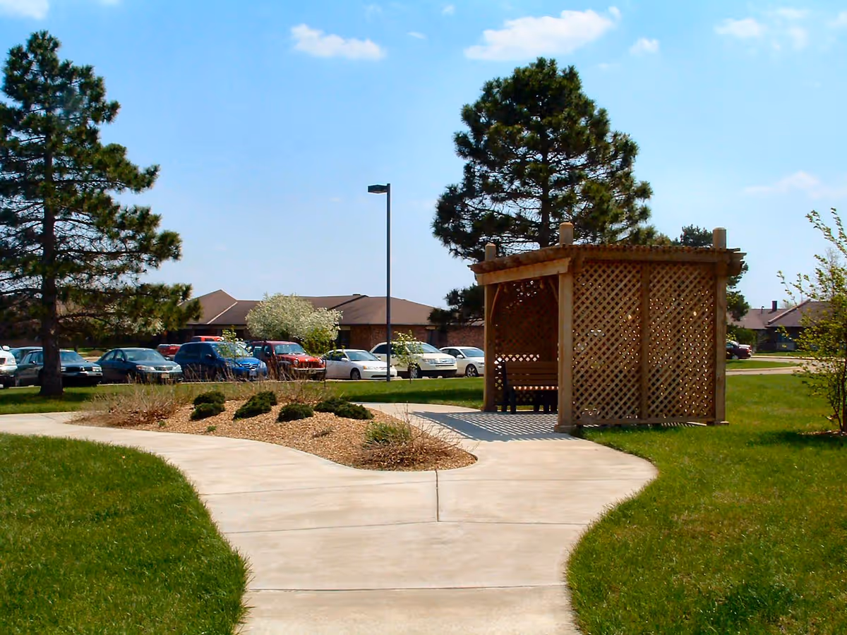 Outdoor scene at Aldersgate Village Life Plan Community featuring a paved walkway that splits into two paths, a wooden lattice gazebo with benches, green grass, trees, parked cars, and residential buildings in the background under a blue sky with some clouds.