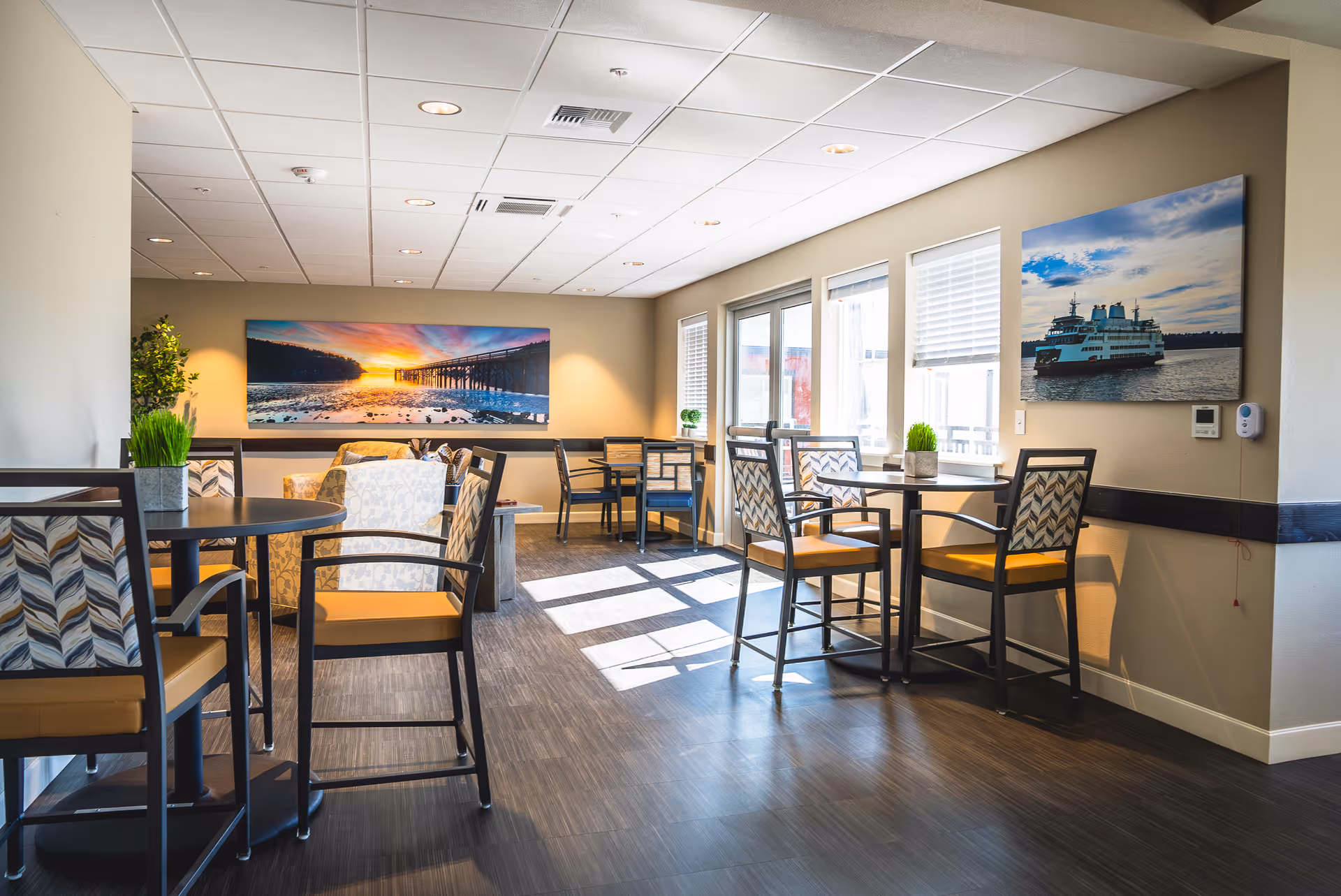 A bright and modern common area with several tables and chairs arranged for seating. The room has large windows letting in natural light, two landscape photographs on the walls, and small potted plants on the tables. The flooring is dark wood, and the ceiling has recessed lighting.