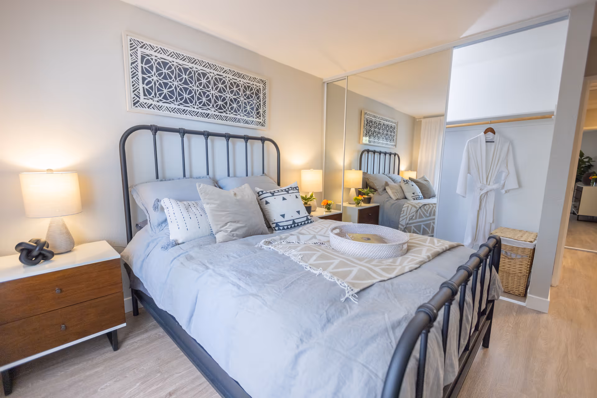 A neatly made bed with a black metal frame in a bedroom. The bed has light gray bedding and several decorative pillows. On the left side of the bed is a wooden nightstand with a lamp and a decorative knot sculpture. Above the bed is a framed geometric art piece. To the right, there is a closet with mirrored sliding doors and a white robe hanging inside. The room has light-colored flooring and walls.