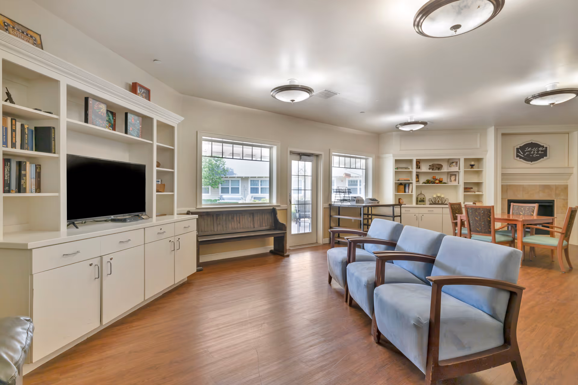A bright and spacious living room area in a senior living facility with wooden flooring, three blue cushioned chairs with wooden armrests, a wooden bench under two windows, a white built-in entertainment center with a TV and shelves containing books and decorative items, a round wooden table with four chairs near a fireplace, and ceiling lights.