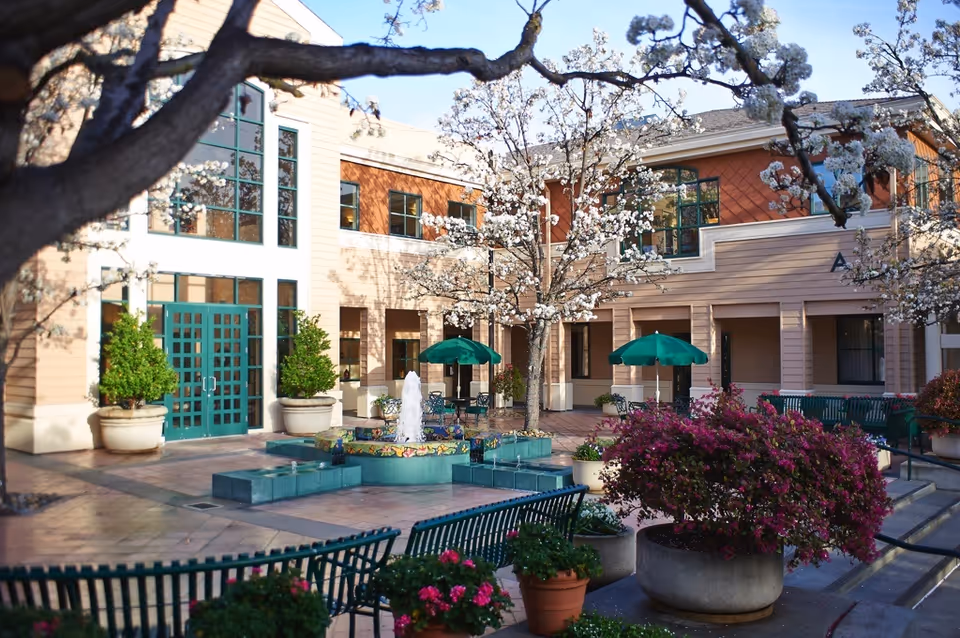 Outdoor courtyard area of The Terraces of Los Gatos featuring blooming trees, a central water fountain, green benches, potted plants with flowers, and green umbrellas over tables. The building exterior is visible with large windows and beige and brown siding.