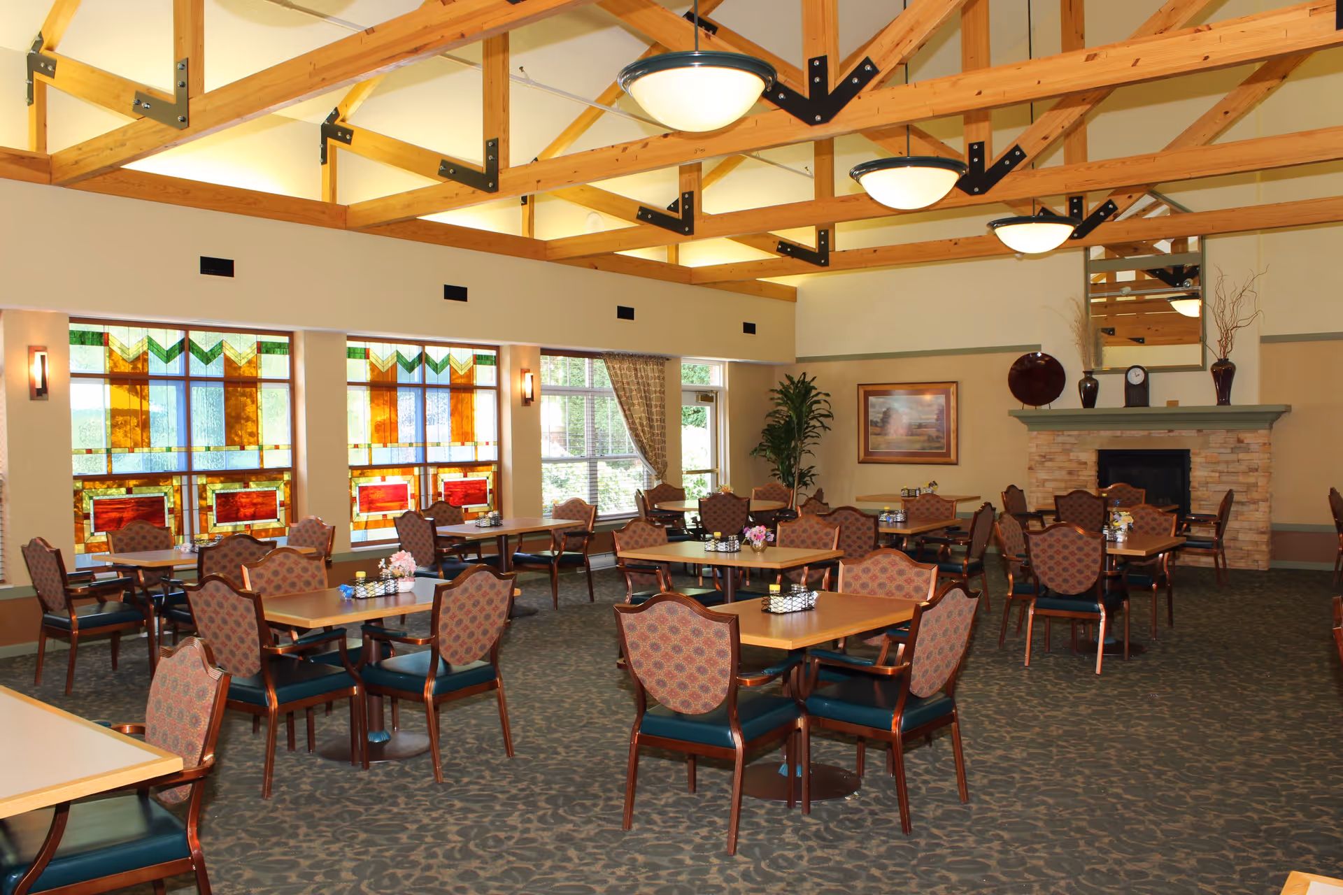 A spacious dining room with multiple wooden tables and cushioned chairs arranged neatly. The room features large stained glass windows on one side, a stone fireplace with decorative items on the mantel, and exposed wooden beams on the ceiling with hanging light fixtures. There is a large plant and framed artwork on the walls, and the carpet has a subtle patterned design.