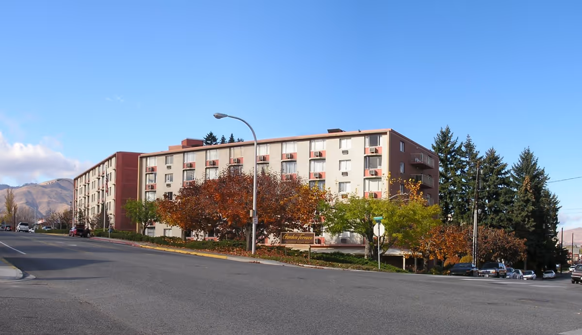 Exterior view of a multi-story retirement home building named Garden Terrace Retirement Home, surrounded by trees with autumn foliage, situated on a street corner with a clear blue sky above.