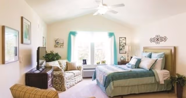 A bright and cozy bedroom with a large bed dressed in teal and beige bedding, a patterned armchair, a TV on a wooden dresser, and a large window with teal curtains letting in natural light.
