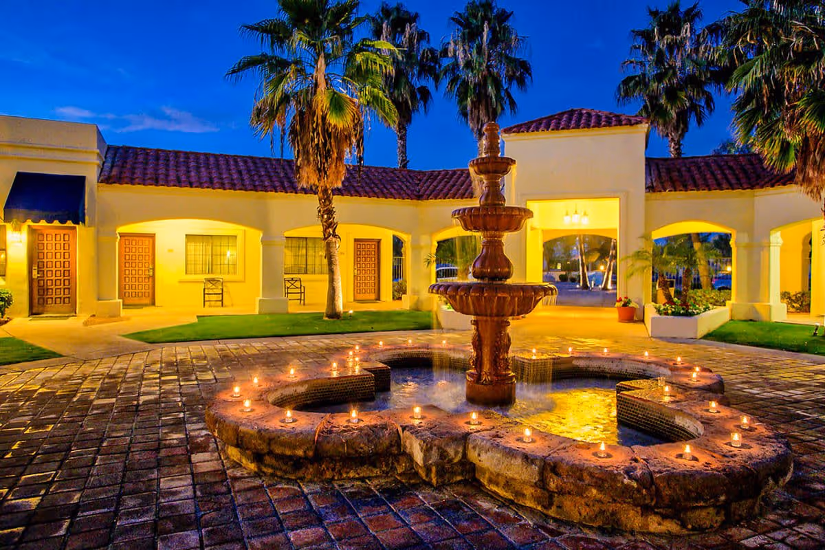 A lit multi-tiered fountain surrounded by candles in a tiled courtyard in front of a Mediterranean-style building with palm trees at dusk.
