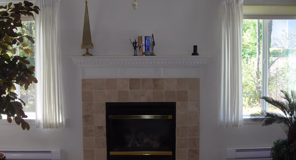 A cozy indoor scene featuring a fireplace with a tiled surround and a white mantel. On the mantel are decorative items including a tall triangular sculpture, two small figurines holding books, and a small black object. The fireplace is flanked by two windows with white curtains, and there are green plants visible near the windows.