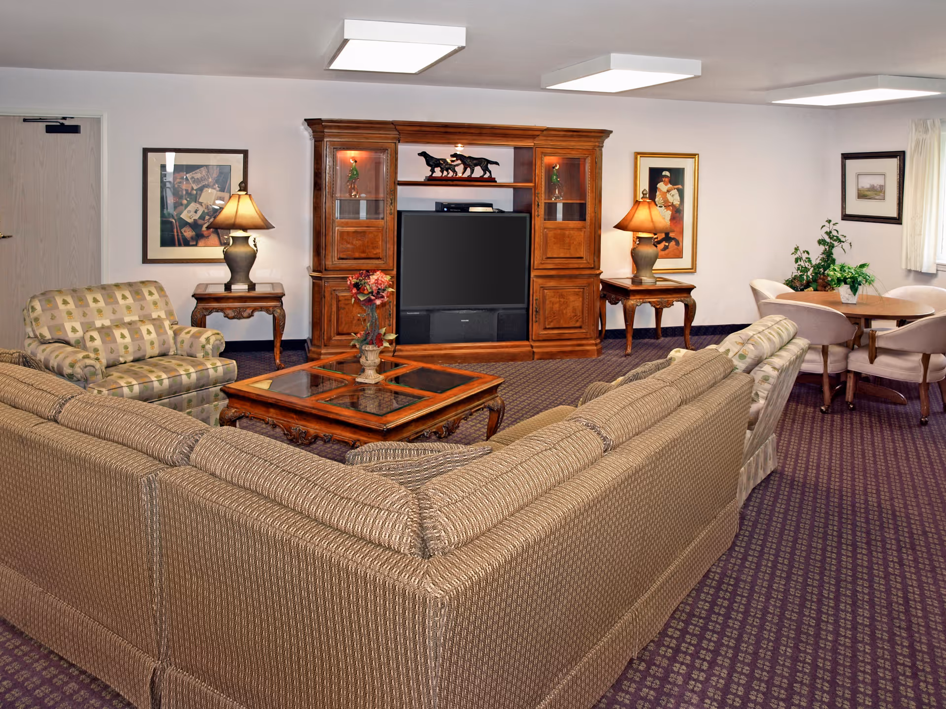 A cozy living room with a large beige sectional sofa, a patterned armchair, and a wooden coffee table with a floral centerpiece. The room features a wooden entertainment center with a TV and decorative items, two side tables with lamps, framed artwork on the walls, and a small round dining table with four chairs near a window with curtains.