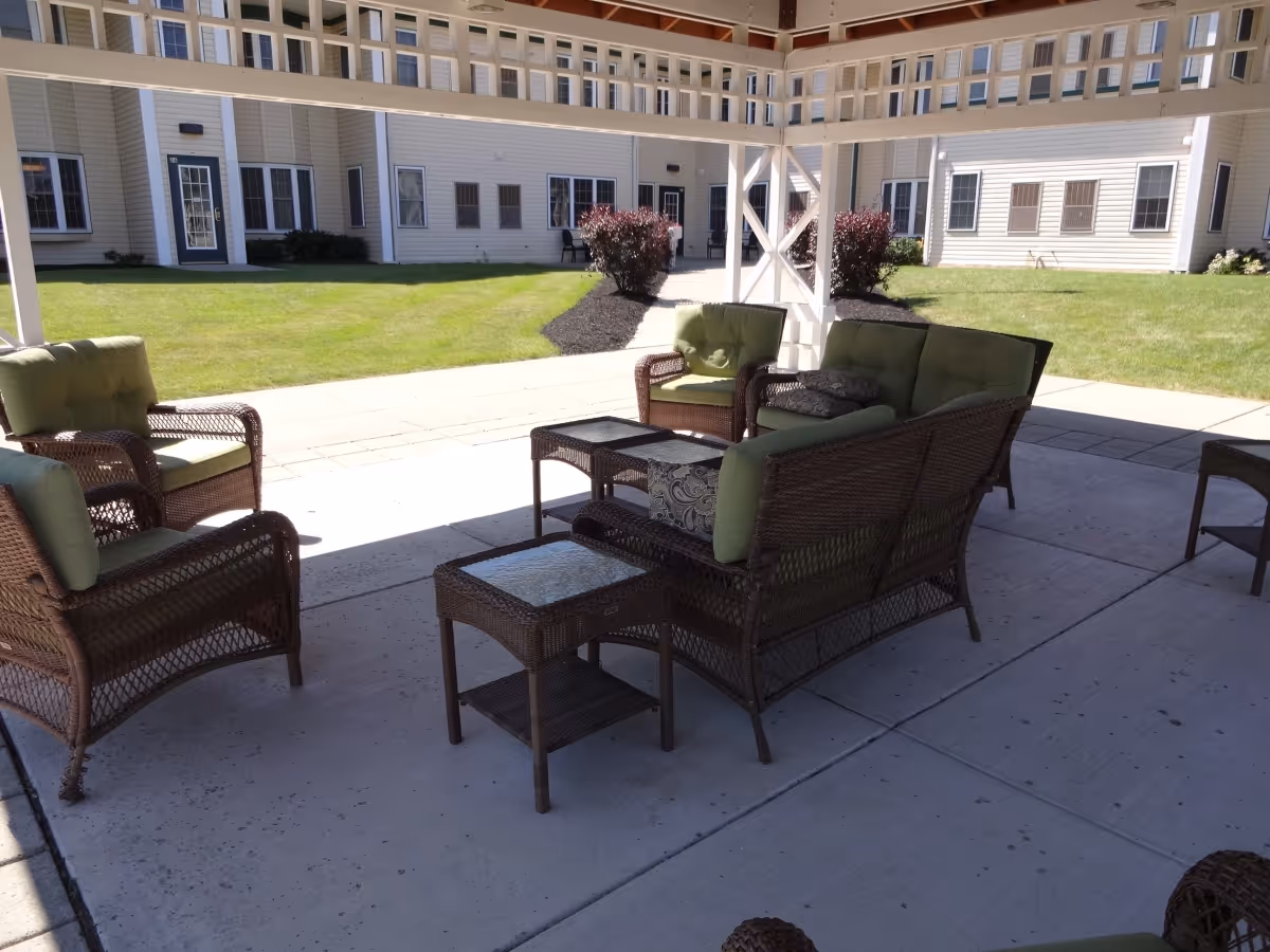 Outdoor patio area with wicker furniture including chairs, a loveseat, and glass-top tables under a pergola. The background shows a well-maintained lawn and the exterior of a light-colored building with multiple windows and doors.