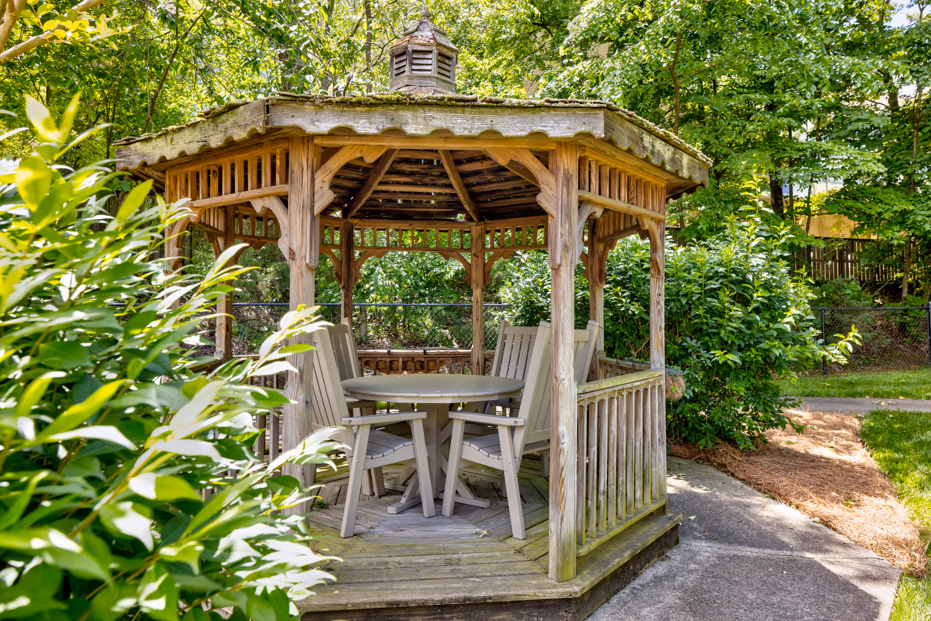 A wooden gazebo with a shingled roof situated in a garden area surrounded by green bushes and trees. Inside the gazebo, there is a round table with four beige chairs arranged around it. A concrete pathway runs beside the gazebo.