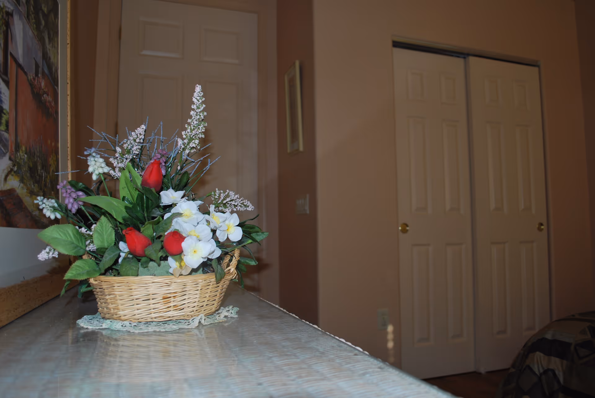 A close-up view of a wicker basket filled with artificial flowers including red roses and white blossoms, placed on a glass-topped table. In the background, there are beige walls, a framed picture, and two closed white doors with gold-colored knobs.