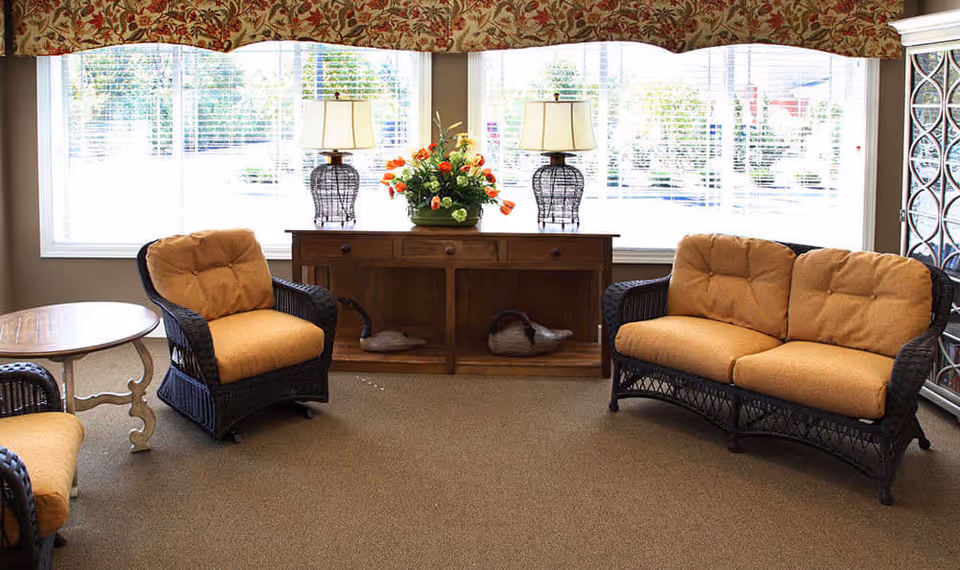 A cozy sitting area with wicker furniture including a loveseat and two armchairs with mustard-colored cushions. A wooden sideboard with two table lamps and a flower arrangement is placed against a large window with white blinds and a floral valance. A round wooden side table is next to one of the armchairs.