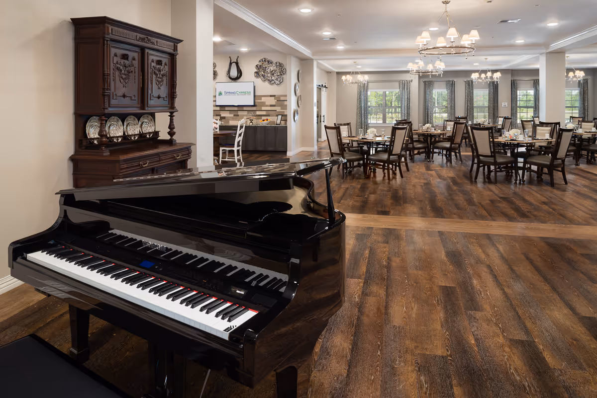 Interior view of a senior living facility dining room with multiple dining tables and chairs arranged neatly. In the foreground, there is a black grand piano. The room has wooden flooring, chandeliers, and large windows with curtains allowing natural light to enter. A wooden cabinet with decorative plates is visible against the wall, and a small desk area with a chair and a screen displaying 'Spring Cypress' is seen in the background.