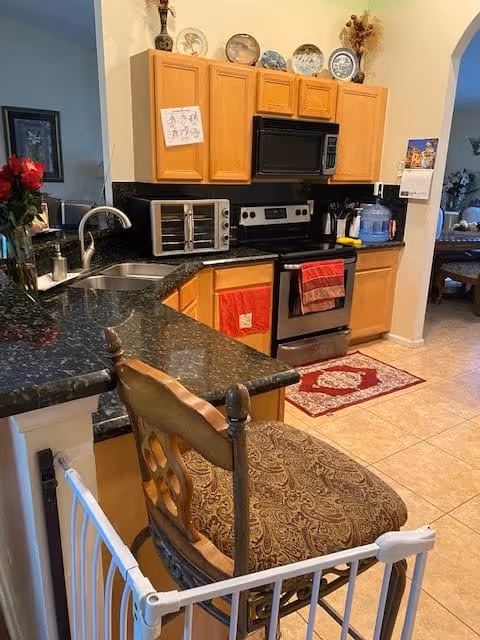 A kitchen with wooden cabinets, black granite countertops, a stainless steel oven and microwave, a toaster oven, and a double sink. There is a patterned cushioned chair at the counter, a red rug on the tiled floor, and various decorative plates displayed above the cabinets. A water dispenser and some kitchen utensils are also visible on the counter.