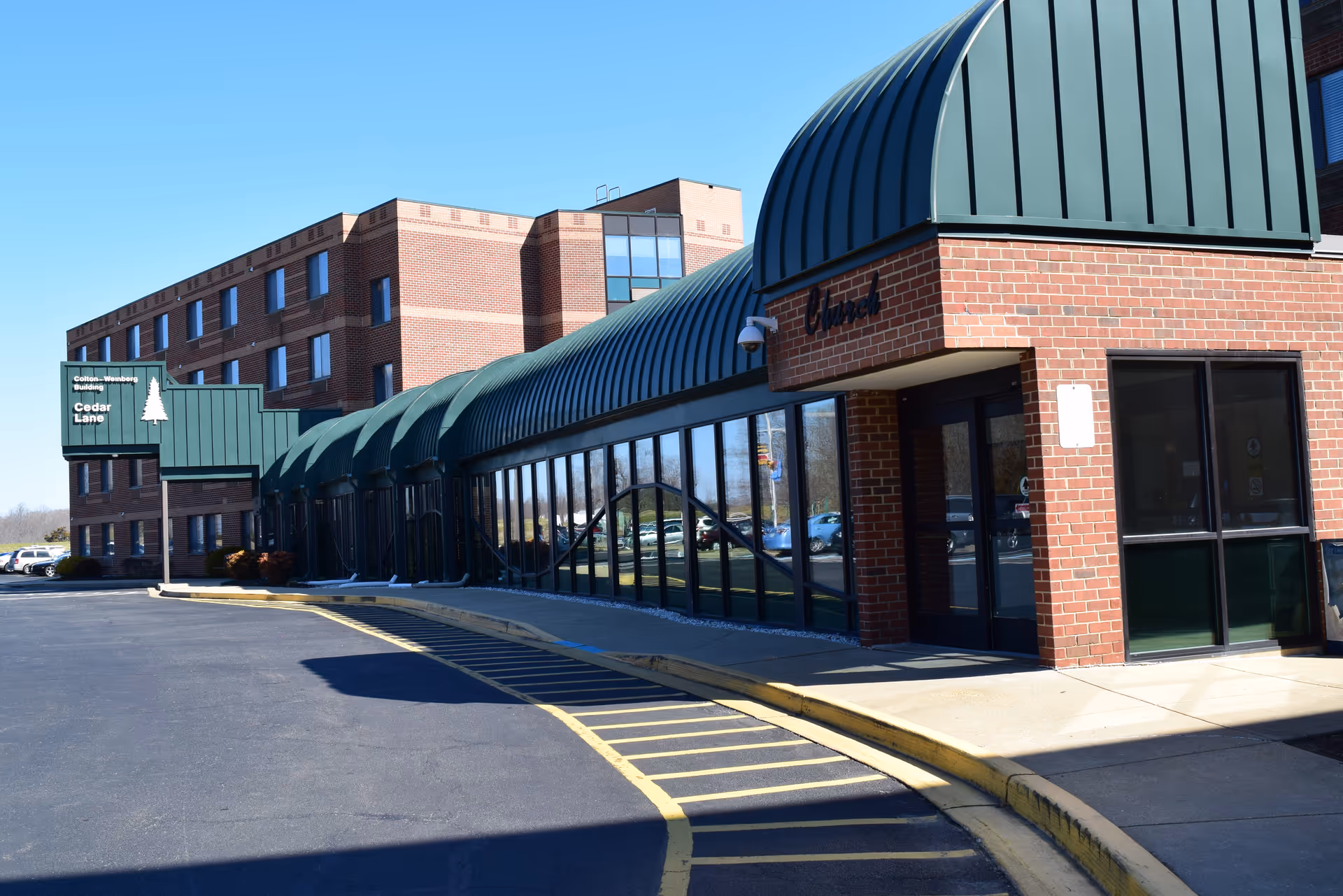Exterior view of Cedar Lane Senior Living Community building with a brick facade and green metal awnings over the entrance and windows. The building has multiple stories and a parking lot in front with yellow painted curbs and lines.