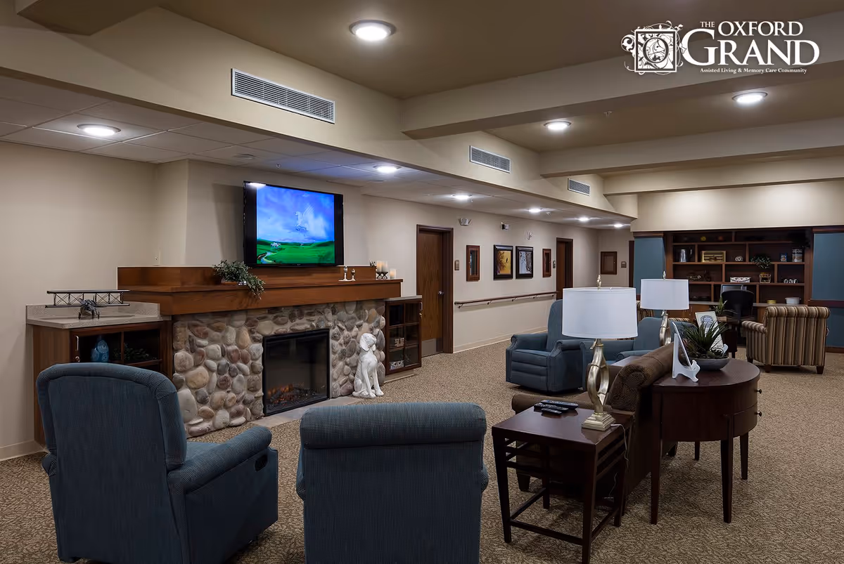 A cozy living room area in The Oxford Grand assisted living facility featuring a stone fireplace with a mounted flat-screen TV above it, two blue upholstered armchairs facing the fireplace, a brown sofa with a side table and lamp, and a wooden bookshelf in the background. The room is well-lit with ceiling lights and decorated with framed pictures on the wall.