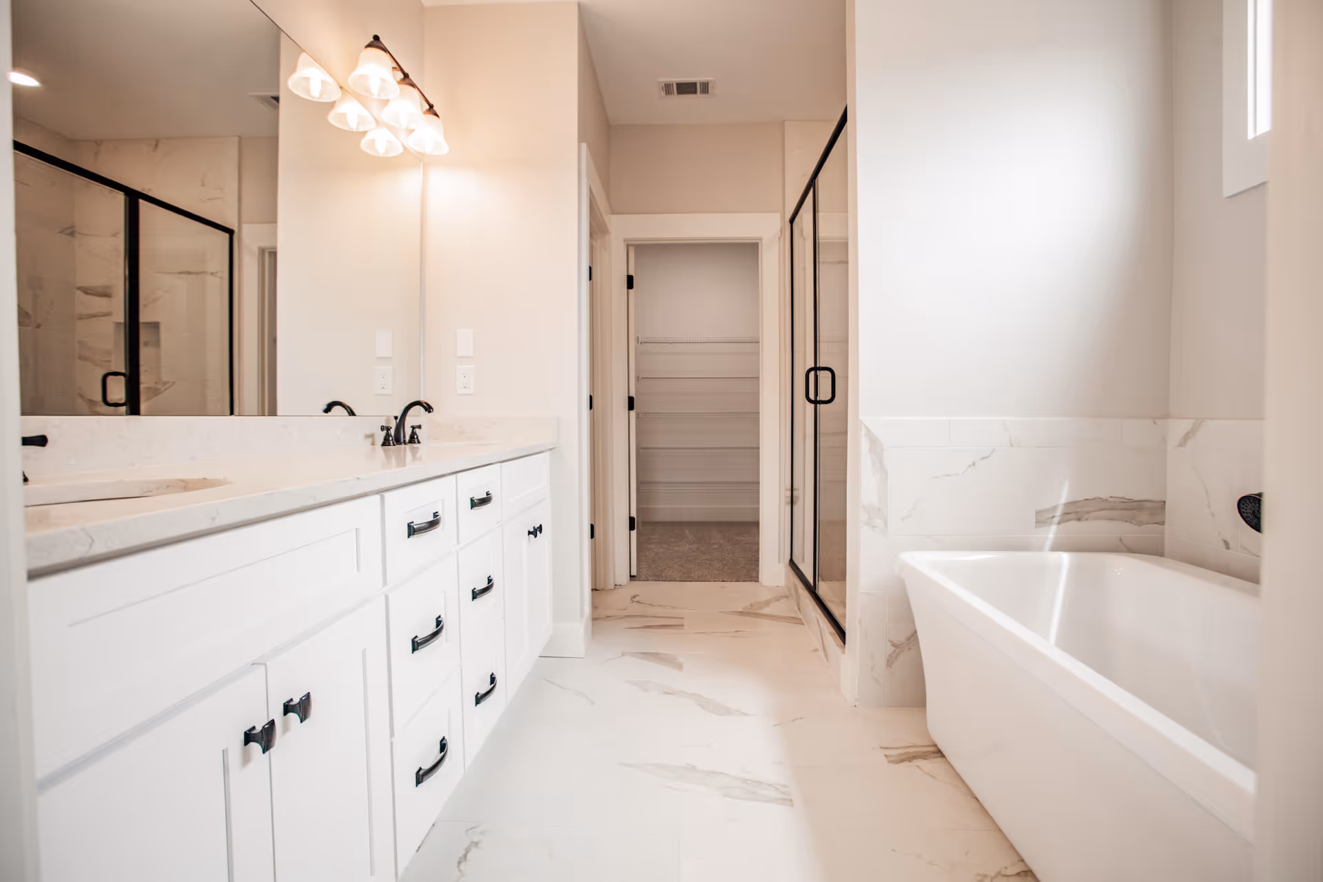 Bright and modern bathroom with white marble flooring and walls, a large white vanity with multiple drawers and black handles, a wide mirror with three light fixtures above it, a glass-enclosed shower with black framing, and a white bathtub next to a window.