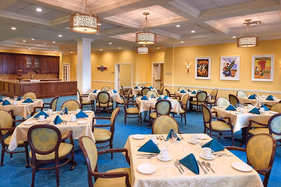 A spacious dining room with multiple round tables covered in cream-colored tablecloths, each set with white plates, cups, silverware, and blue folded napkins. The room features yellow walls, blue carpet, decorative ceiling lights, and framed vintage posters on the walls. A wooden counter and cabinetry are visible in the background.