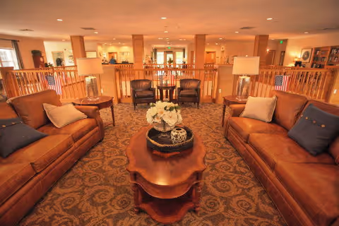 A spacious and warmly lit living room area with two brown leather sofas facing each other, each adorned with pillows. Between them is a wooden coffee table with a decorative tray and a flower arrangement. Behind the sofas are two armchairs with a small table in between. The room features wooden railings and a patterned carpet, with soft lighting from table lamps and recessed ceiling lights.