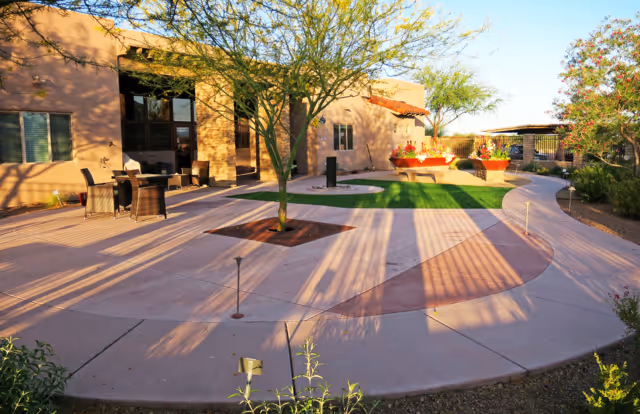 Outdoor patio area at a senior living facility with a curved concrete walkway, green grass patches, trees, potted plants, and outdoor seating including a table with chairs. The building exterior is visible with windows and a shaded entrance.