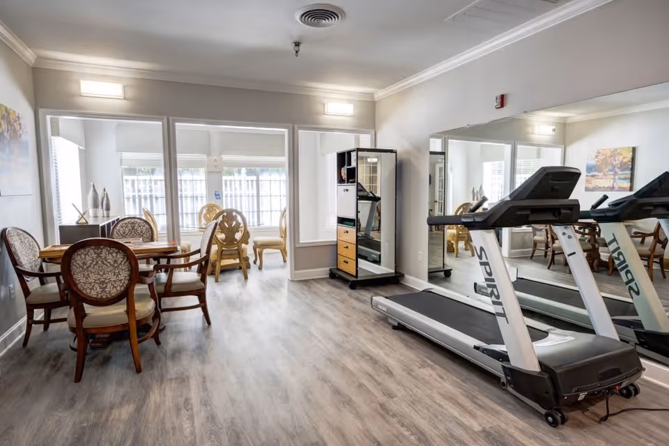 A bright room with a treadmill on the right side in front of a large wall mirror. On the left side, there is a wooden table with four upholstered chairs. In the background, there are large windows letting in natural light and additional chairs near the windows. The room has light-colored walls and wood flooring.