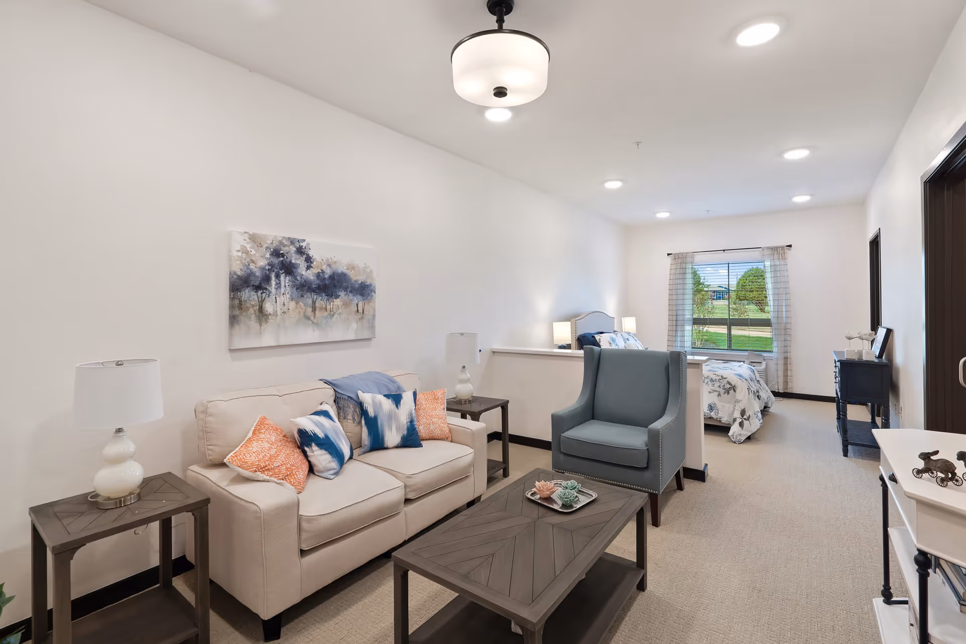 A bright and neatly arranged senior living apartment featuring a beige sofa with colorful cushions, a blue armchair, a wooden coffee table with decorative items, and a bed in the background near a window with curtains. The room has white walls, recessed ceiling lights, and side tables with lamps.