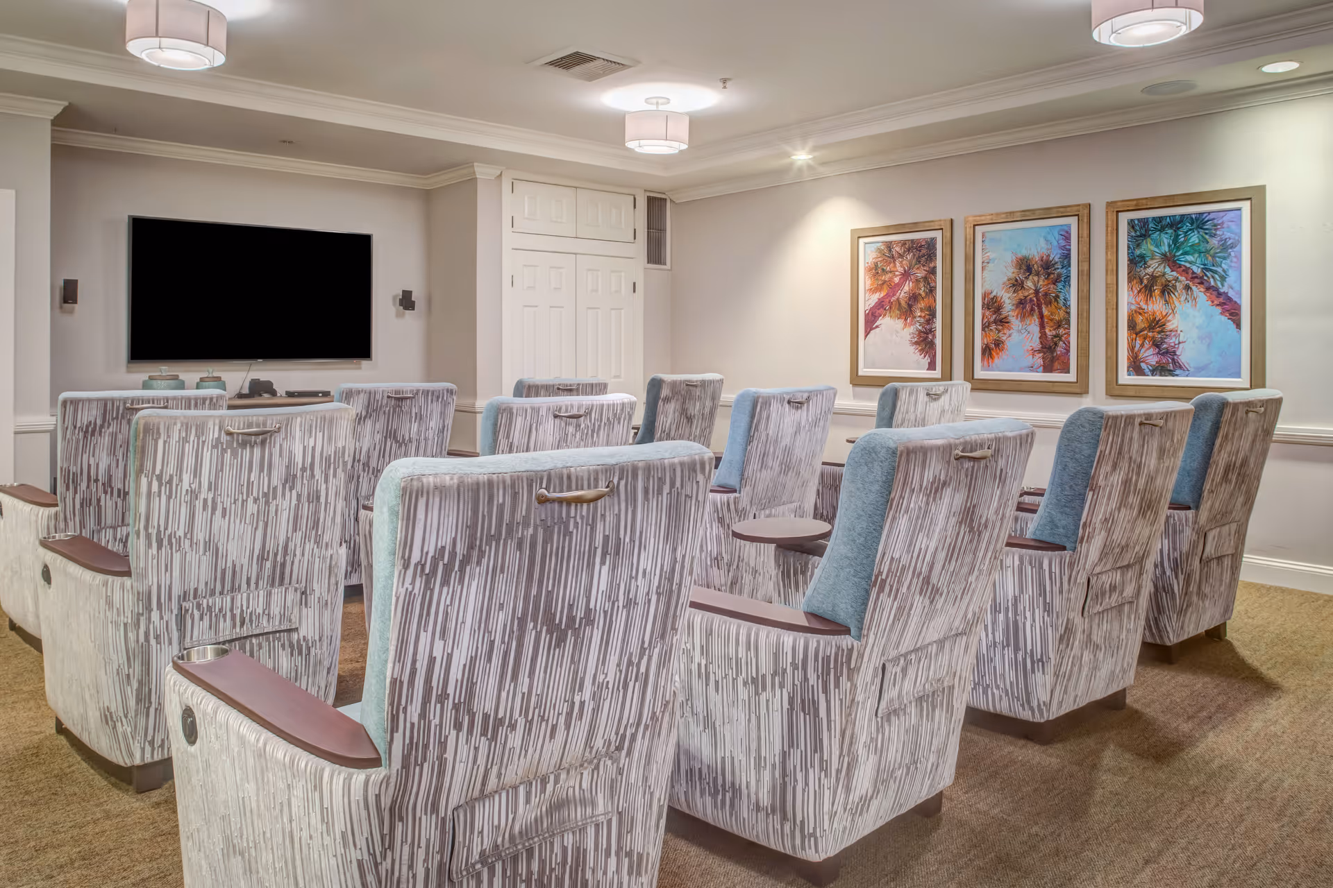 A small theater room with two rows of patterned upholstered chairs facing a large flat-screen TV mounted on the wall. The room has beige walls with three framed pictures of palm trees on the right wall, carpeted floor, and ceiling lights.