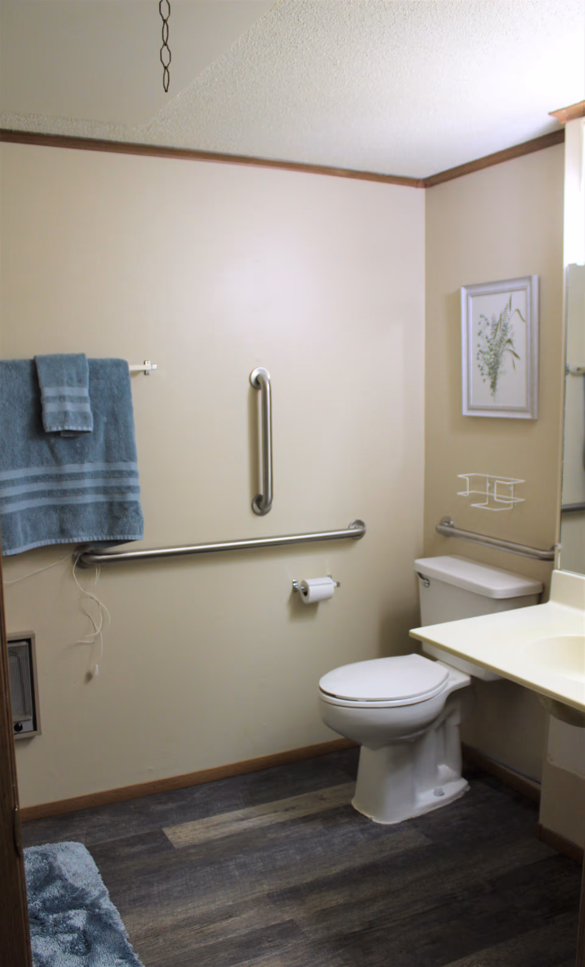 A bathroom with beige walls and wood trim, featuring a white toilet, a white sink with a countertop, a towel rack with blue towels, and metal grab bars on the walls for support. There is a framed botanical picture hanging above the toilet and a blue bath mat on the floor.