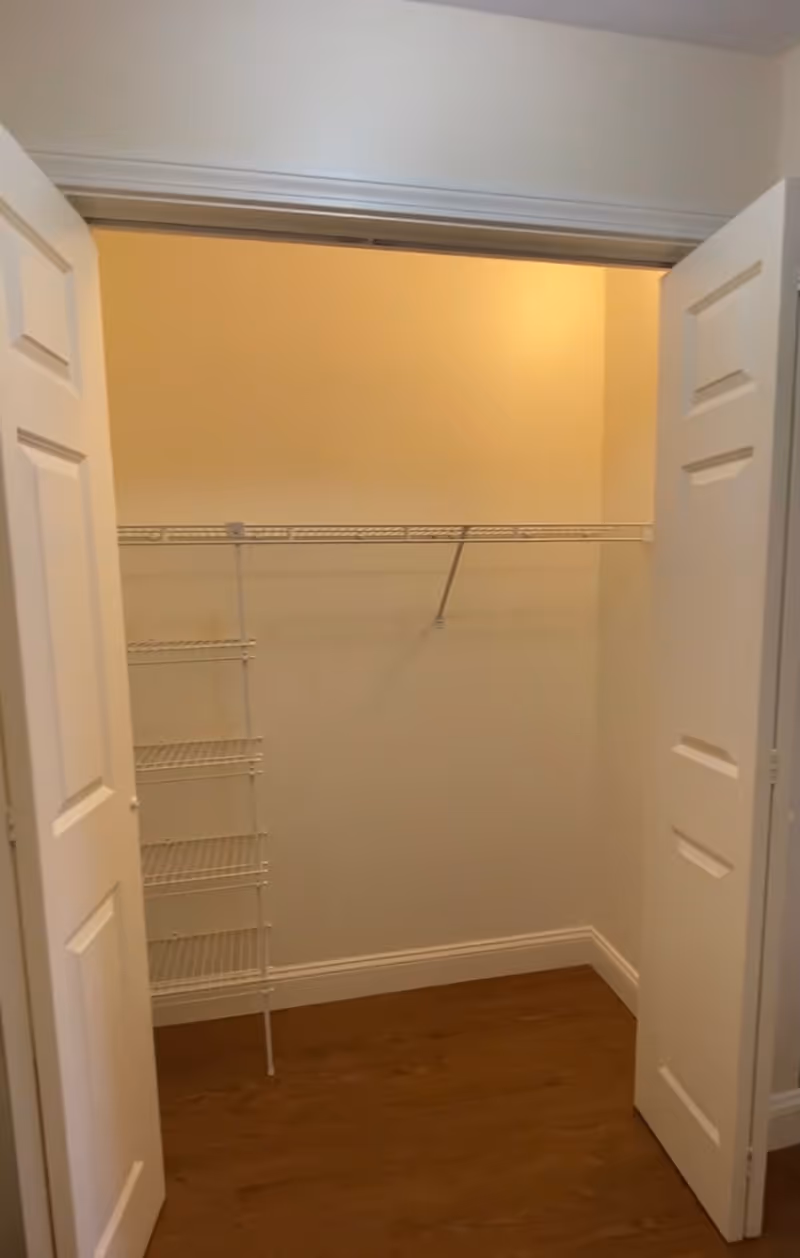 Empty walk-in closet with white double doors, white wire shelving on the left side and back wall, and wooden floor.