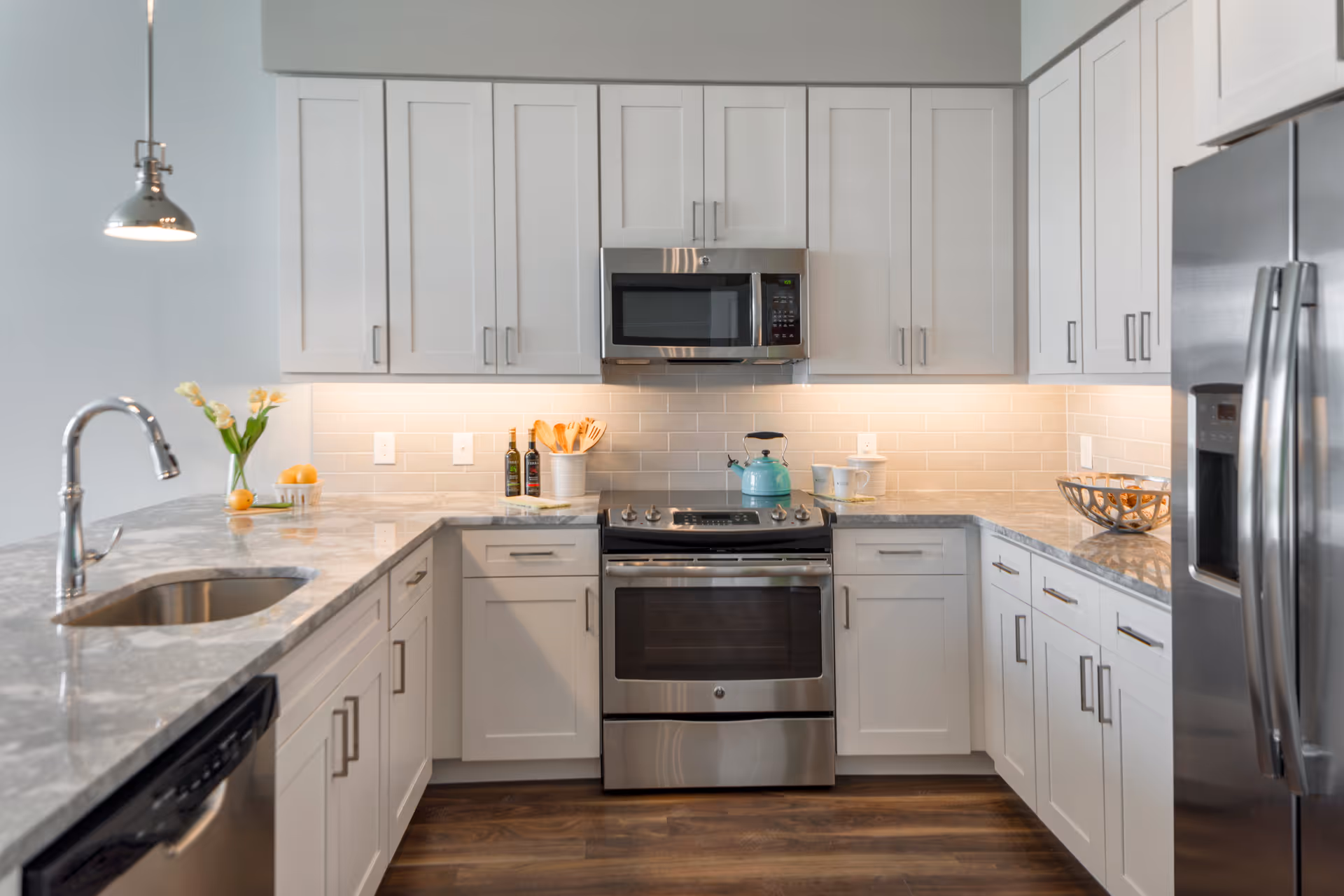 Modern kitchen with white cabinets, stainless steel appliances including a refrigerator, oven, and microwave. Marble countertops with a sink and faucet on the left side, a teal kettle on the stove, and decorative items such as a vase with flowers, olive oil bottles, and a fruit bowl on the counters.