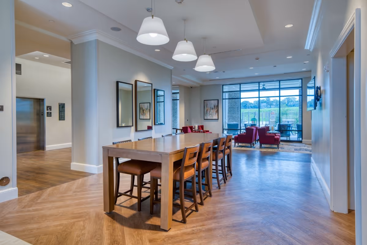 A spacious interior common area with a long wooden table surrounded by chairs in the foreground. The room has wooden flooring, three pendant lights hanging above the table, and mirrors on the wall. In the background, there are red armchairs near large windows that provide a view of an outdoor patio and greenery beyond.