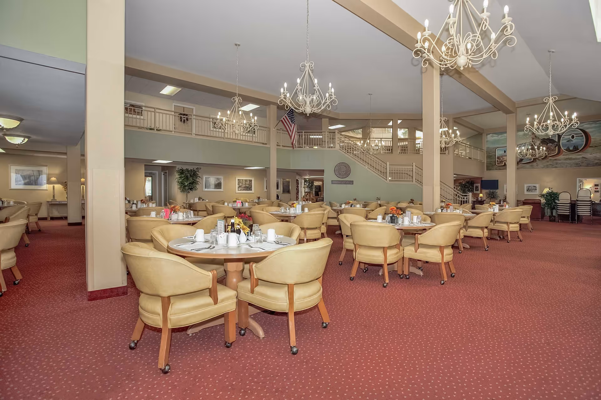 Spacious senior dining room with round tables set for meals, cushioned chairs, chandeliers, and a second-floor mezzanine.