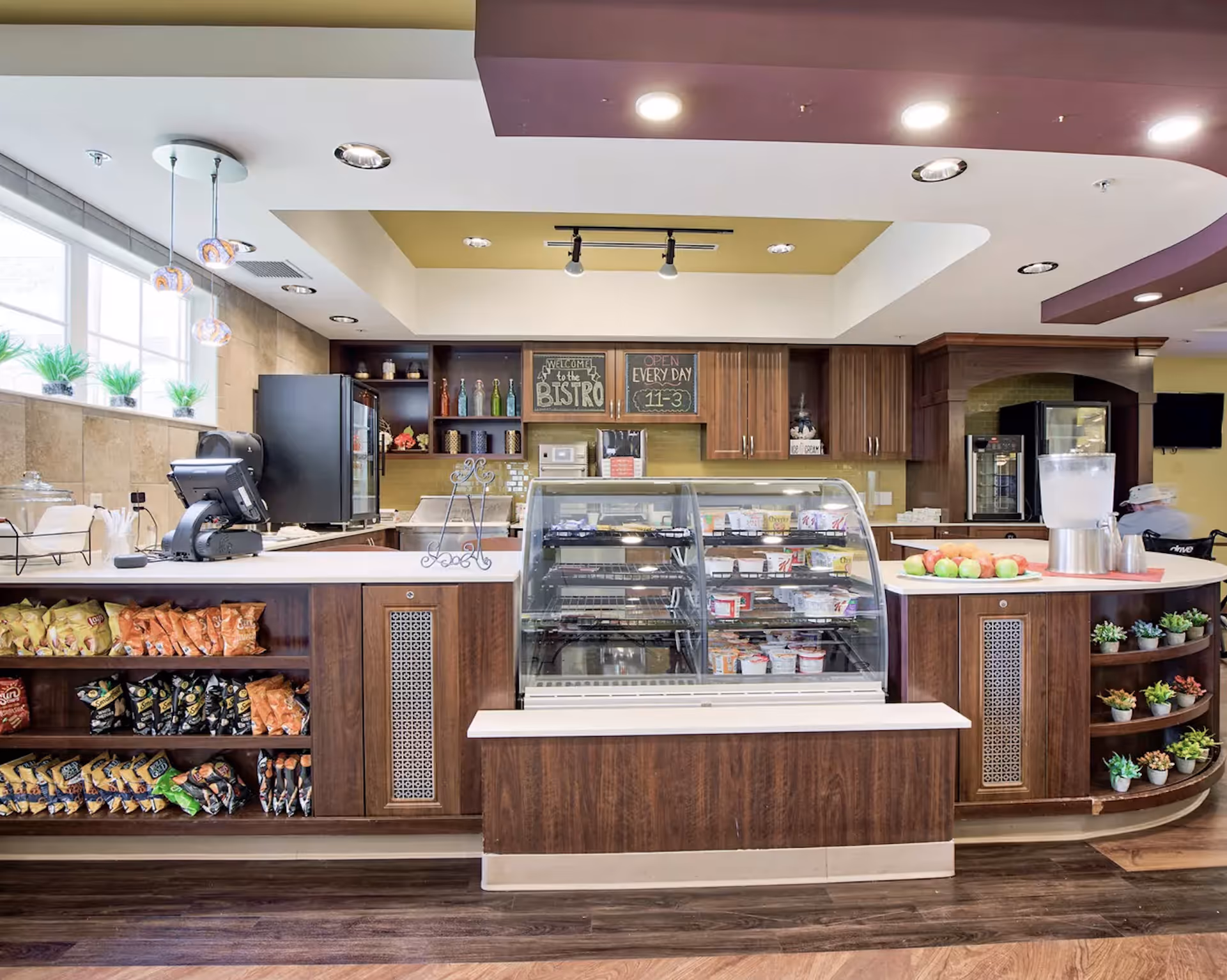 Interior view of a bistro-style snack and beverage counter with a glass display case containing various snacks and food items. The counter has shelves stocked with bags of chips and a section with small potted plants. Behind the counter are wooden cabinets, a refrigerator, and a sign that reads 'Welcome to the Bistro' and 'Open Every Day 11-3'. The area is well-lit with ceiling lights and pendant lights near a window with small plants on the sill.