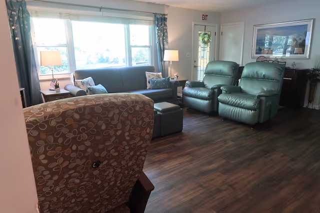 A cozy living room with a large window letting in natural light. The room features a blue sofa with decorative pillows, two green recliner chairs, a patterned armchair in the foreground, a small ottoman, and two side tables with lamps. There is a door with a wreath and a framed picture on the wall.