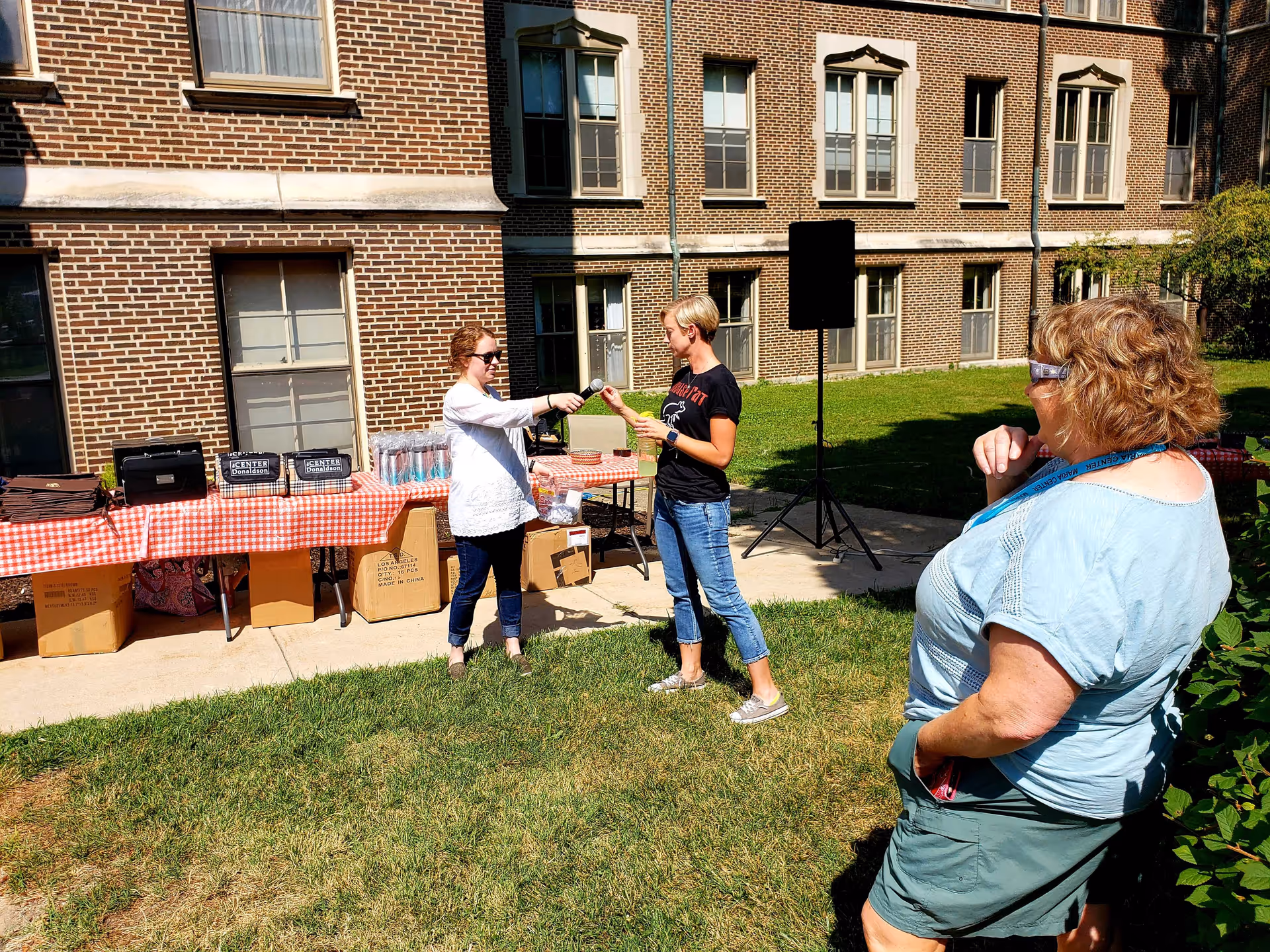Three women outdoors near a brick building. One woman in a white top and sunglasses is handing a microphone to another woman in a black t-shirt and jeans. A third woman in a light blue top and shorts is standing nearby watching. There is a table with a red and white checkered tablecloth and boxes underneath it in the background.