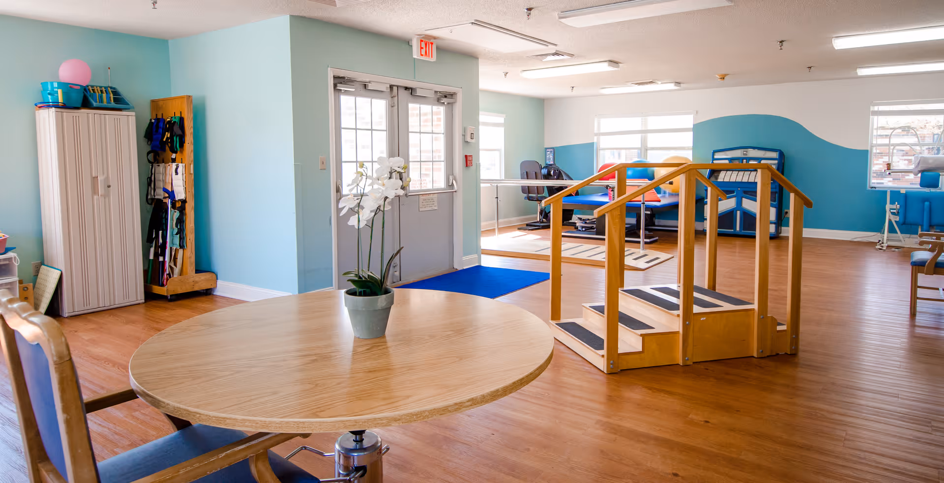 A bright and spacious therapy room with light blue walls and wooden flooring. In the foreground, there is a round wooden table with a potted white orchid on top and a chair with blue upholstery. In the middle of the room, there is a wooden set of stairs with handrails on both sides, likely used for physical therapy exercises. The background shows exercise equipment, including a treadmill and other therapy apparatus near large windows letting in natural light. A double door with an exit sign above it is visible on the left side of the room.