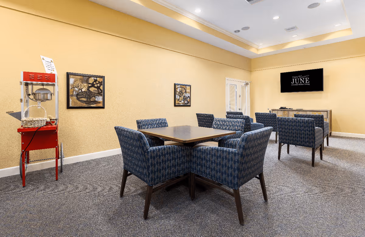 A cozy common area with a square table surrounded by four blue patterned chairs in the foreground and two blue patterned armchairs facing a wall-mounted TV in the background. The walls are painted yellow, and there is a red popcorn machine on the left side. The carpet is gray, and the ceiling has recessed lighting.