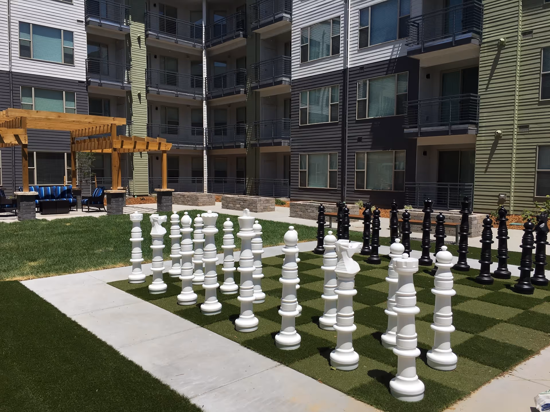 Outdoor courtyard area of a residential facility with a large chessboard featuring oversized black and white chess pieces on a grass-like surface. Surrounding the chessboard are paved walkways, a wooden pergola with blue cushioned seating underneath, and multi-story apartment buildings with balconies and windows in the background.