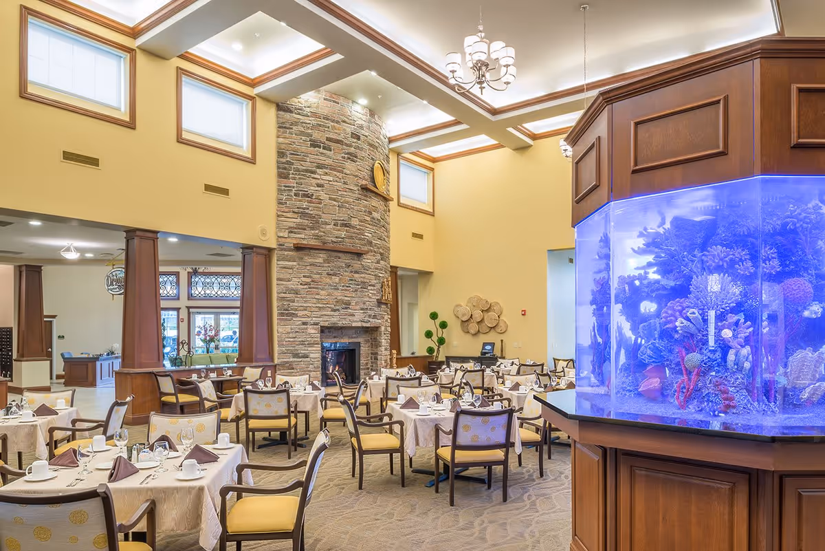 A spacious dining room in a senior living facility with multiple tables covered in white tablecloths, each set with cups, glasses, and napkins. The room features a tall stone fireplace, high ceilings with recessed lighting, and large windows near the ceiling. On the right side, there is a large, illuminated aquarium with colorful coral and fish. The decor includes wooden pillars and a chandelier hanging from the ceiling.